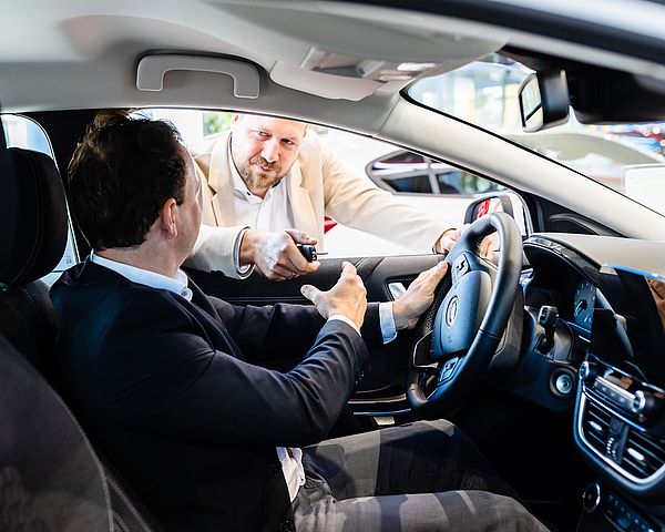 Dunkelhaariger Mann sitzt in Auto und unterhält sich mit blondem Mann durch das Fahrerfenster.