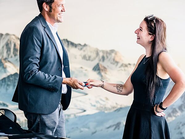 Dunkelhaariger Mann und dunkelhaarige Frau stehen neben einem Auto und übergeben sich einen Autoschlüssel.