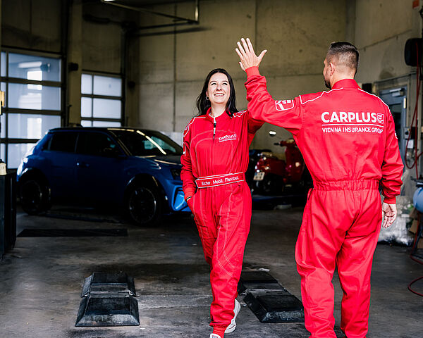 Zwei Mitarbeitende von CARPLUS in roten Arbeitsanzügen geben sich in einer Kfz-Werkstatt ein High-Five. Im Hintergrund stehen mehrere Fahrzeuge.