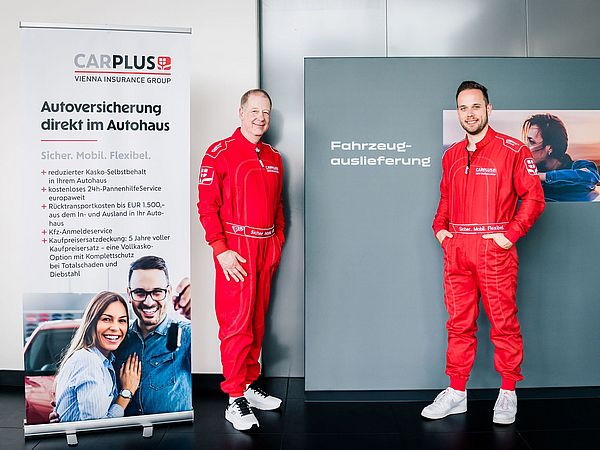 Zwei Männer in roten Rennanzuügen stehen vor einer Werbetafel von carplus.