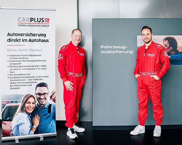 Zwei Männer in roten Rennanzuügen stehen vor einer Werbetafel von carplus.