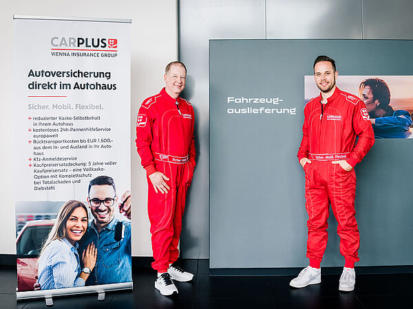 Zwei Männer in roten Rennanzuügen stehen vor einer Werbetafel von carplus.