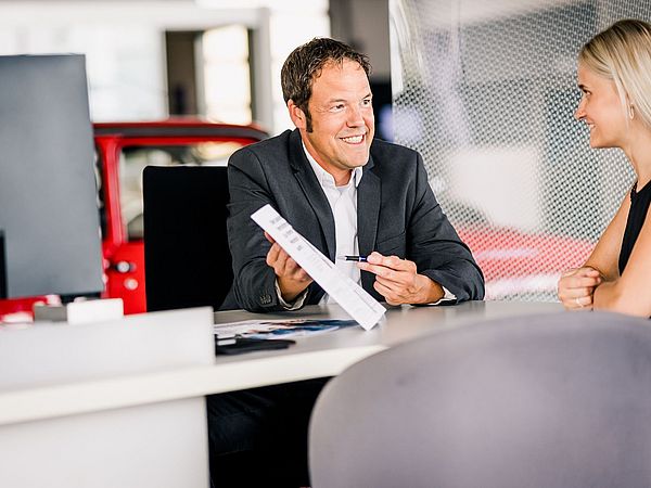 Mann mit Jacket sitzt an einem Schreibtisch und hält Dokumente in der Hand die er einen jungen blonden Dame zeigt.