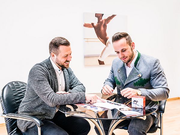 Zwei dunkelhaarige Männer sitzen an einem kleinen Besprechungstisch und schauen sich Unterlagen an.