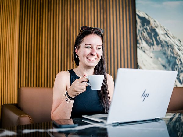 Junge, dunkelhaarige Dame sitzt mit Kaffeetasse vor dem Laptop.