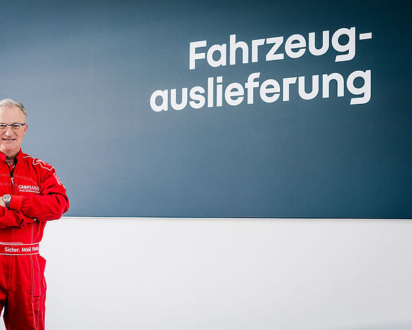 Ein Mann mit grauen Haaren und Brille steht mit verschränkten Armen in rotem Rennanzug.