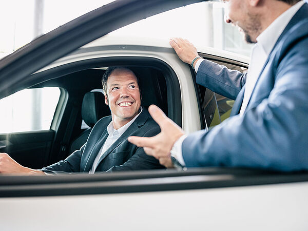 Mann mit Jacket sitzt lächelnd im Auto. Ein anderer Mann erklärt ihm etwas.