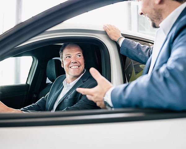 Mann mit Jacket sitzt lächelnd im Auto. Ein anderer Mann erklärt ihm etwas.