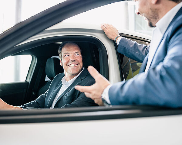 Mann mit Jacket sitzt lächelnd im Auto. Ein anderer Mann erklärt ihm etwas.