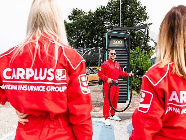 Mann steht bei Elektro-Aufladestation und hält den Stecker in der Hand, 2 Damen sieht man von hinten im roten Rennanzug mit Carplus-Logo am Rücken.