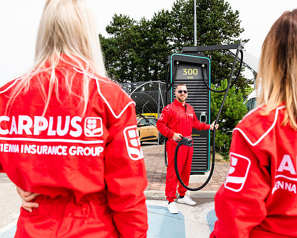 Mann steht bei Elektro-Aufladestation und hält den Stecker in der Hand, 2 Damen sieht man von hinten im roten Rennanzug mit Carplus-Logo am Rücken.