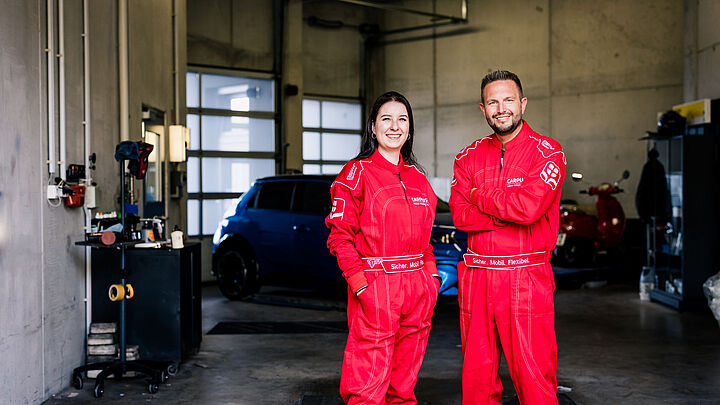 Eine junge Frau und ein junger Mann in roten Rennanzügen stehen vor einer Autowerkstatt.
