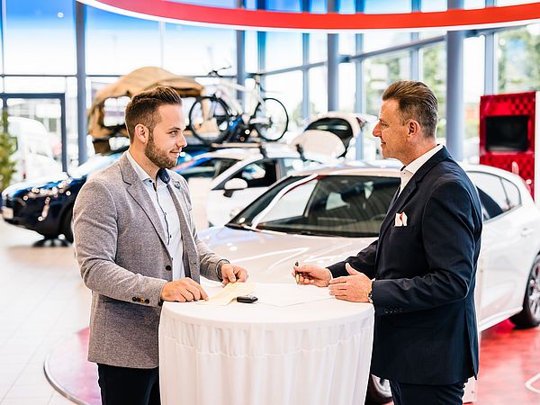 Zwei dunkelhaarige Männer stehen an einem kleinen Besprechungstisch in einem Autohaus.